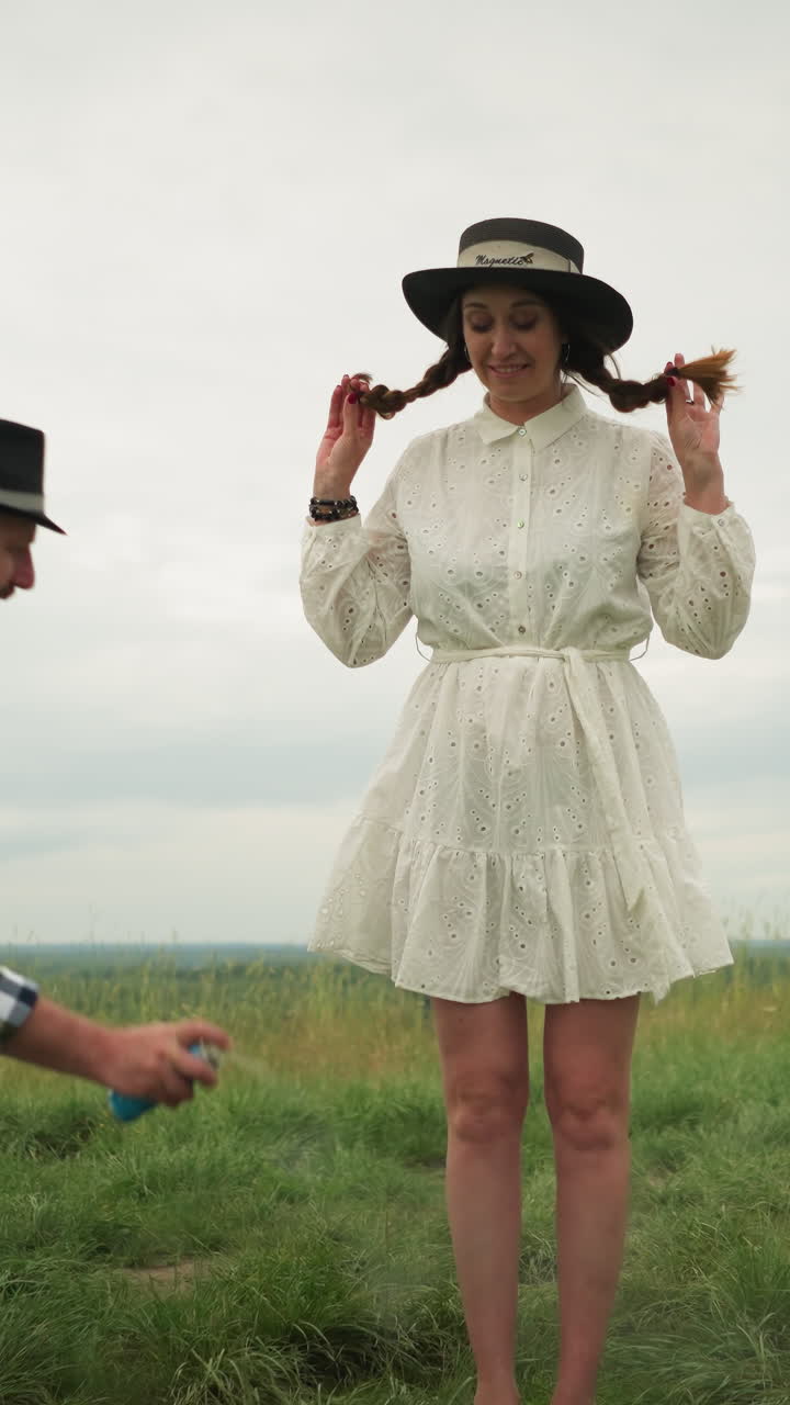 se ve a un hombre con una camisa a cuadros rociando repelente de mosquitos en las piernas de una mujer, que está en un elegante sombrero y un vestido blanco. se paran en un campo de hierba con un lienzo pintado y suministros de arte cerca