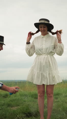 se ve a un hombre con una camisa a cuadros rociando repelente de mosquitos en las piernas de una mujer, que está en un elegante sombrero y un vestido blanco. se paran en un campo de hierba con un lienzo pintado y suministros de arte cerca