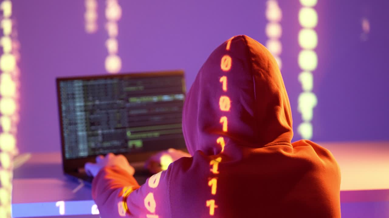 A person in an orange hoodie sits at a laptop, illuminated by streams of orange binary code in a neon purple room, symbolizing hacking, data flow, and digital warfare in cyberspace.