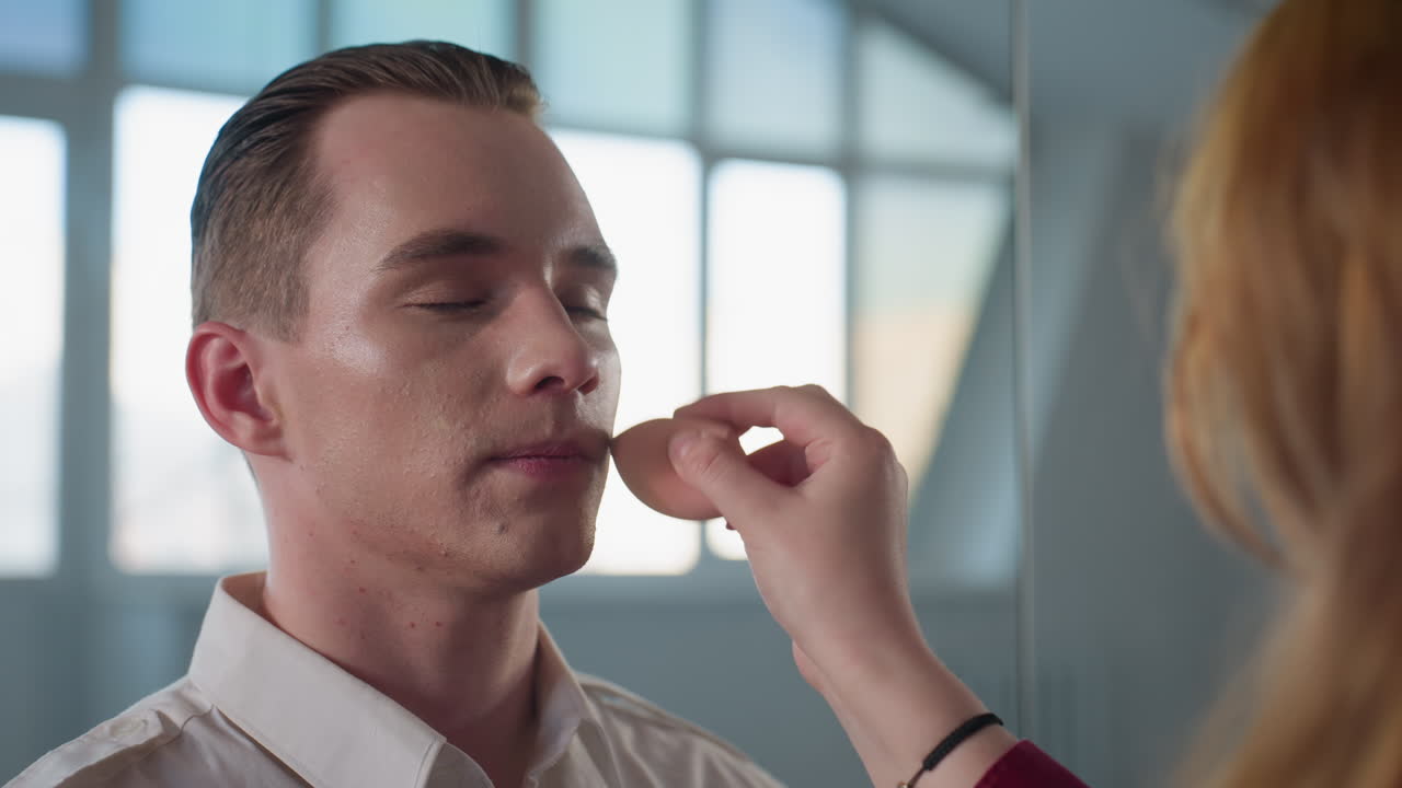 makeup artist blending liquid foundation on model face with sponge in modern studio setting while mirror reflects window light and room details creating professional beauty preparation scene