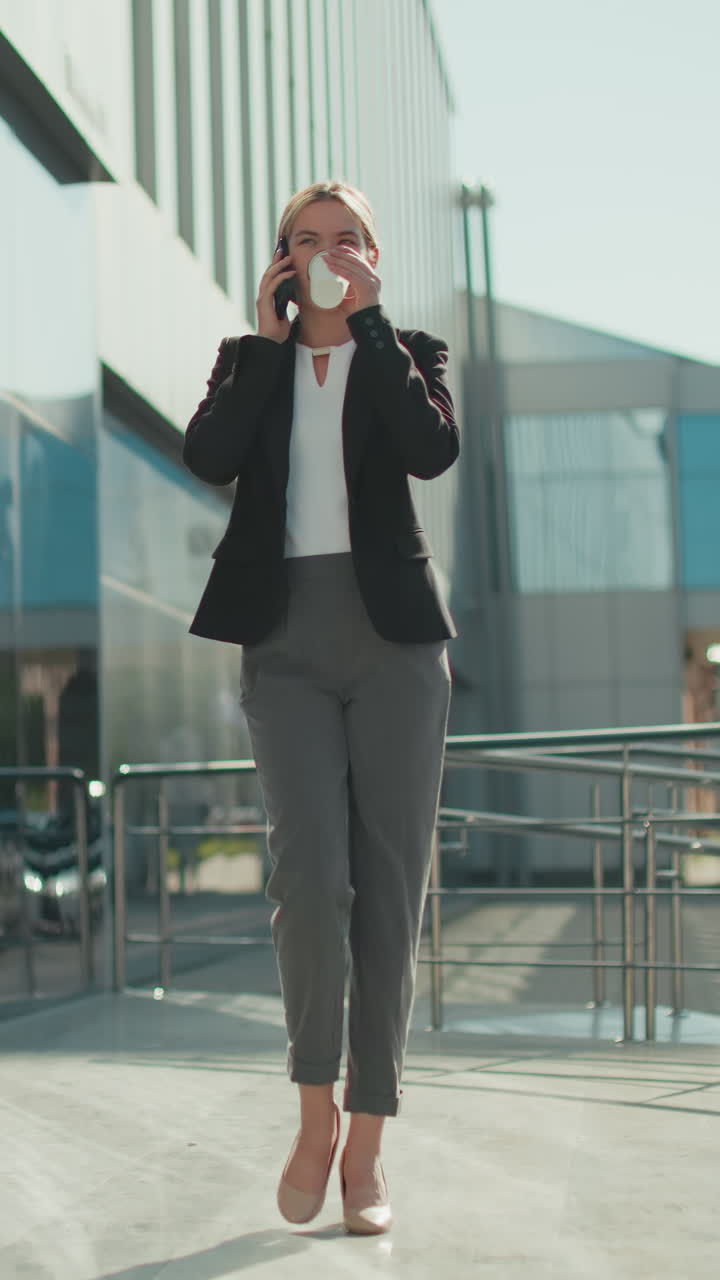 Elegant lady walking with coffee in hand, warm smile as she sips drink during phone call, iron railing surrounds walkway with background featuring parked cars and blurred pedestrian in sunlight