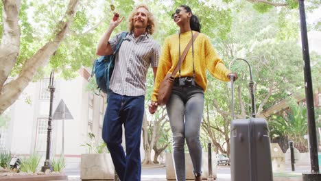 happy diverse couple walking with luggage on sunny day in city