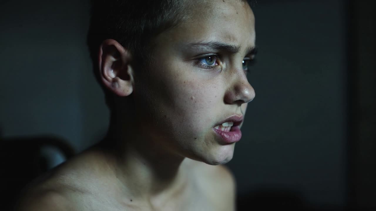 Adolescente che urla per poi calmarsi, con espressione mutevole in una stanza fioca, a torso nudo, elaborando la rabbia, testiera del letto a sinistra.