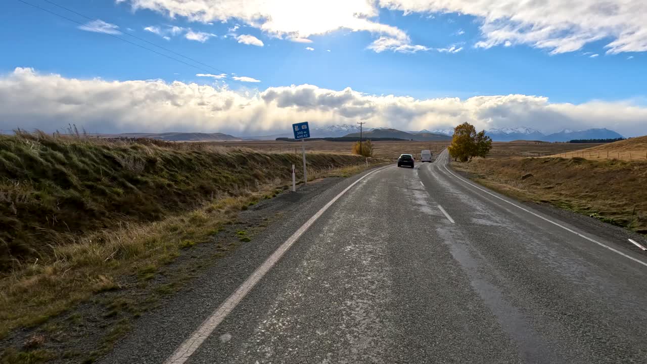 A camper van travels along a wet rural road through open countryside, under dramatic clouds and bright autumn sunlight, captured with smooth forward camera movement