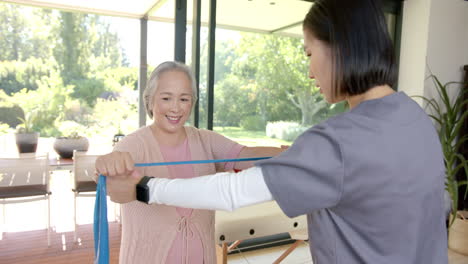 senior asian woman exercising with resistance band, assisted by physiotherapist at home