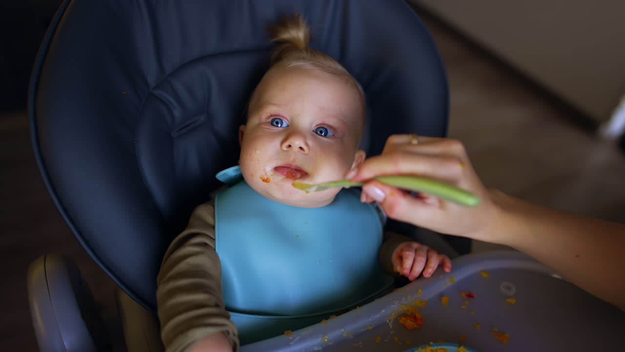 Little kid sitting in high chair looks up at camera in surprise. Mom's hand feeding a baby from spoon. Top view.