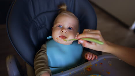 little kid sitting in high chair looks up at camera in surprise. mom's hand feeding a baby from spoon. top view.