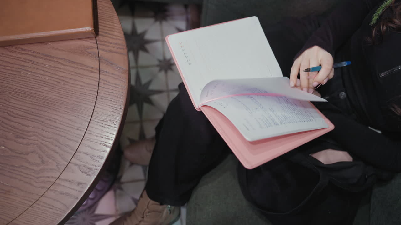 Woman wearing black top sits with legs crossed holding pink notebook and blue pen, gently placing brown notebook on wooden table while preparing to write, captured in a calm thoughtful indoor moment