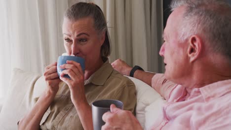 caucasian senior couple drinking coffee cup while talking to each other sitting on the couch at home