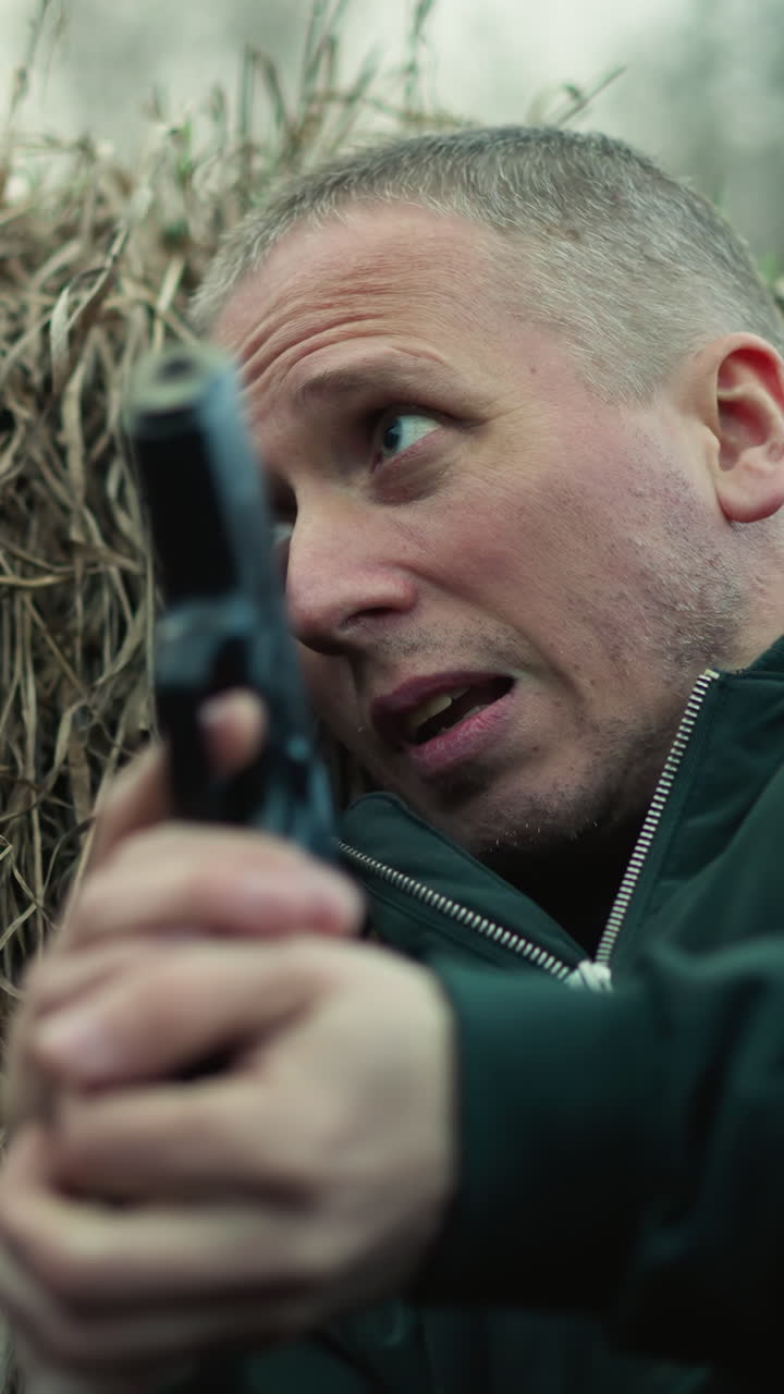 una vista de cerca de un hombre con una chaqueta verde acostado contra la hierba seca, sosteniendo una pistola, y mirando tenso