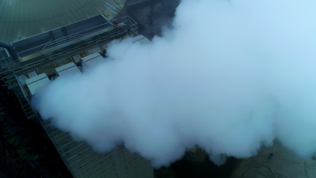 Working premises at elevator plant, thick pipes letting out heavy thick smoke. Flying above the tops of granary silo at the backdrop of grey weather.