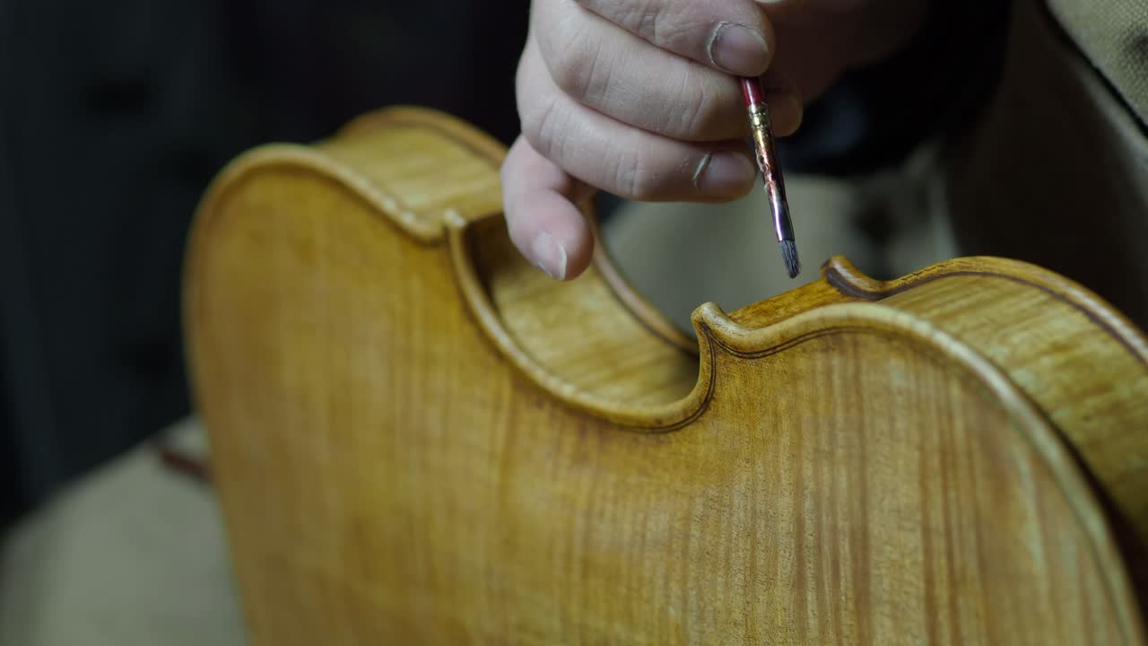 Close-up of a craftsman’s steady hand expertly applying antiquing varnish with a fine paintbrush and finger tamponing to the center bout ribs and edge of a violin, creating authentic aged appearance