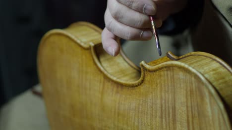 close-up of a craftsman’s steady hand expertly applying antiquing varnish with a fine paintbrush and finger tamponing to the center bout ribs and edge of a violin, creating authentic aged appearance