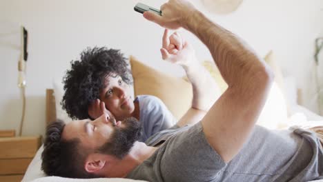 Video-De-Una-Feliz-Pareja-Diversa-Relajándose-En-Casa,-Acostada-En-La-Cama-Usando-Un-Teléfono-Inteligente