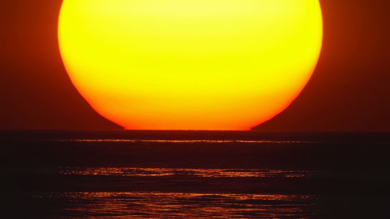 Sol enorme se pondo no horizonte do oceano, luz dourada refletindo em ondas suaves, composição em close-up de ângulo baixo