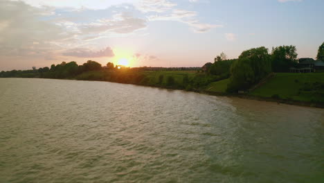 Die-Drohne-Erhebt-Sich-über-Dem-Wasser,-Um-Felder-Und-Kleine-Hüttenbaustellen-Zu-Enthüllen,-Während-Die-Sonne-Im-Hintergrund-Untergeht