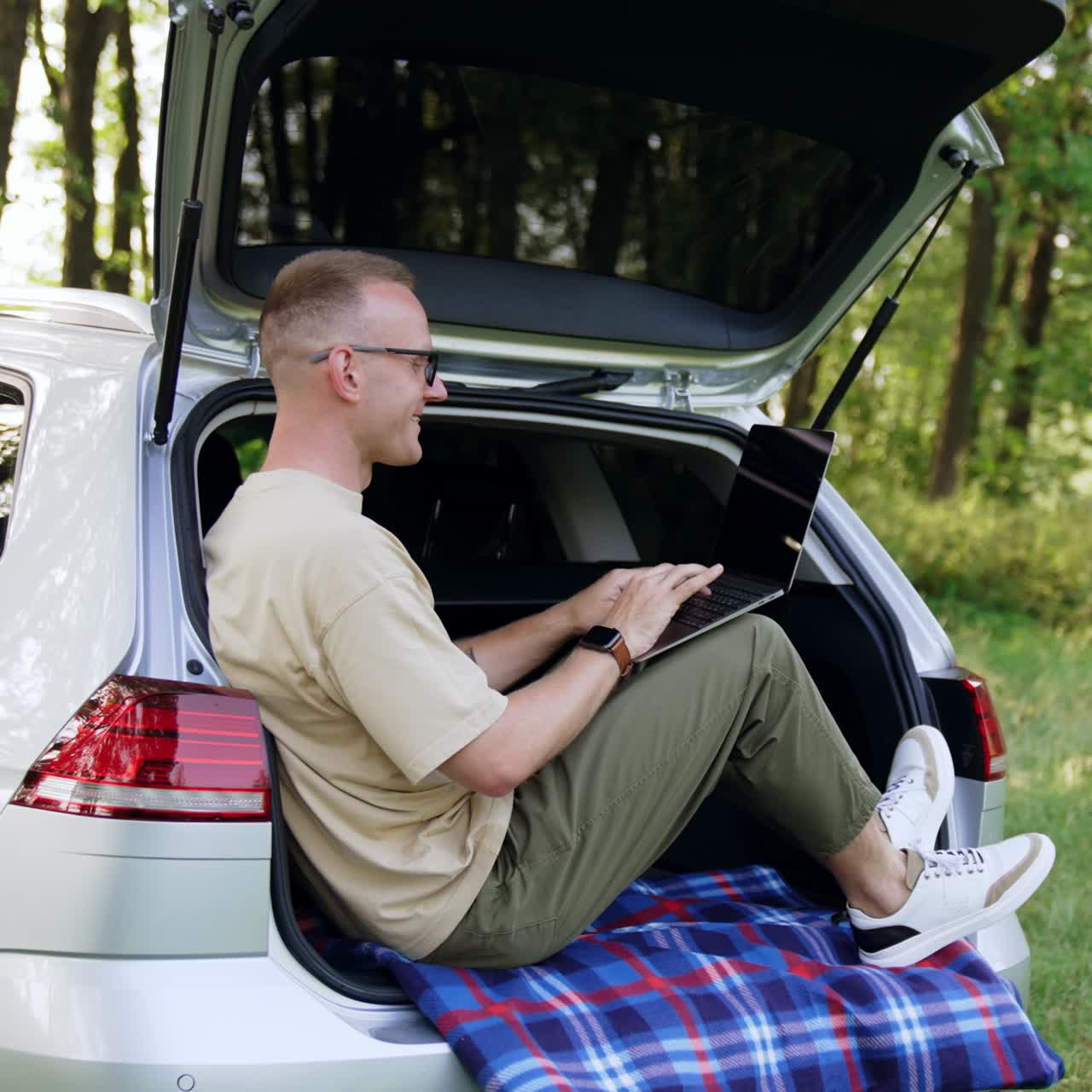 Young male sitting comfortably in his car trunk. Caucasian man working remote on his laptop