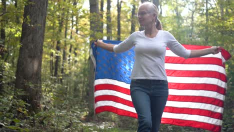 Blonde-Frau-Blickt-Zum-Himmel,-Hält-Eine-Flagge-Hinter-Sich-Und-Hebt-Sie,-Während-Sie-Durch-Einen-Wald-Geht