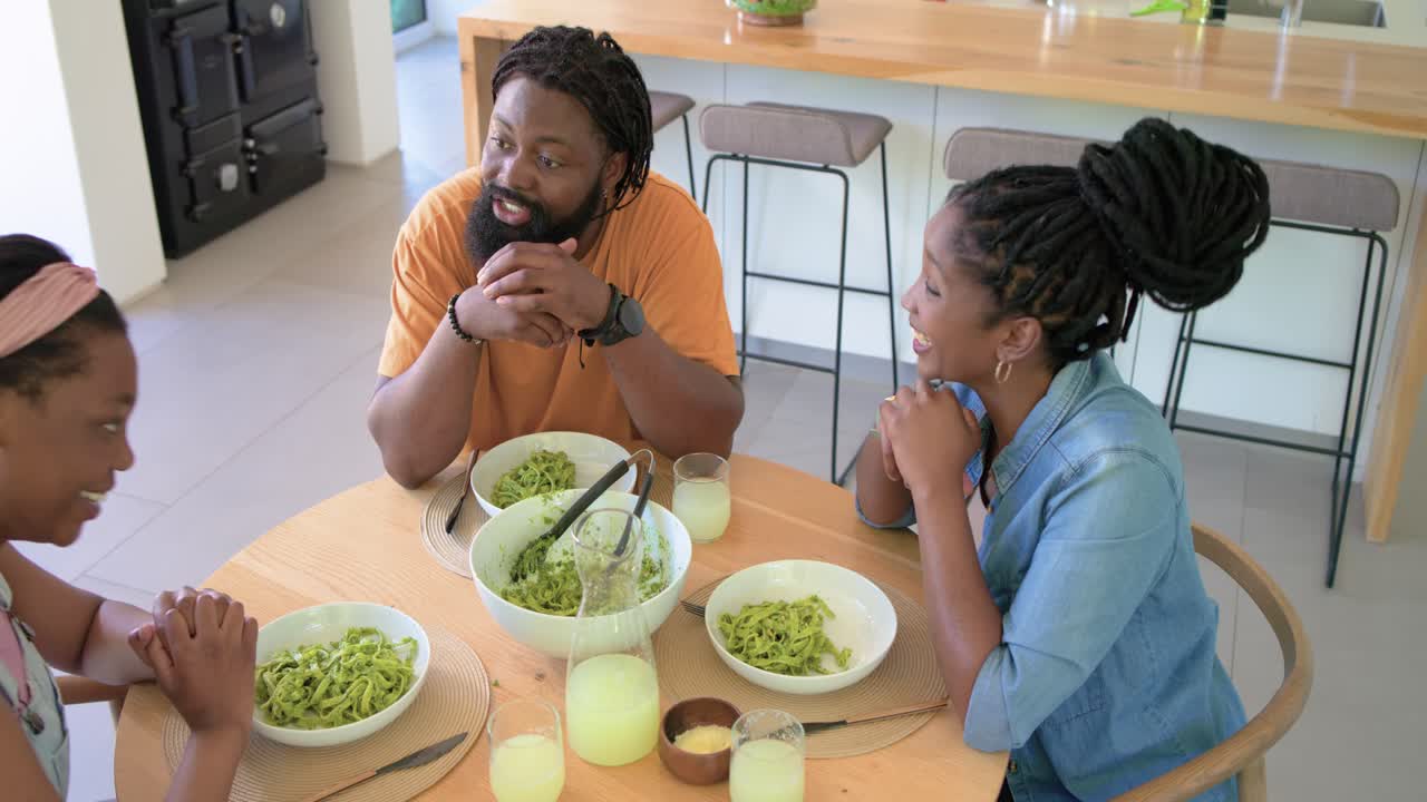 African family talking, then woman wearing denim shirt serving green pasta with tongs in kitchen