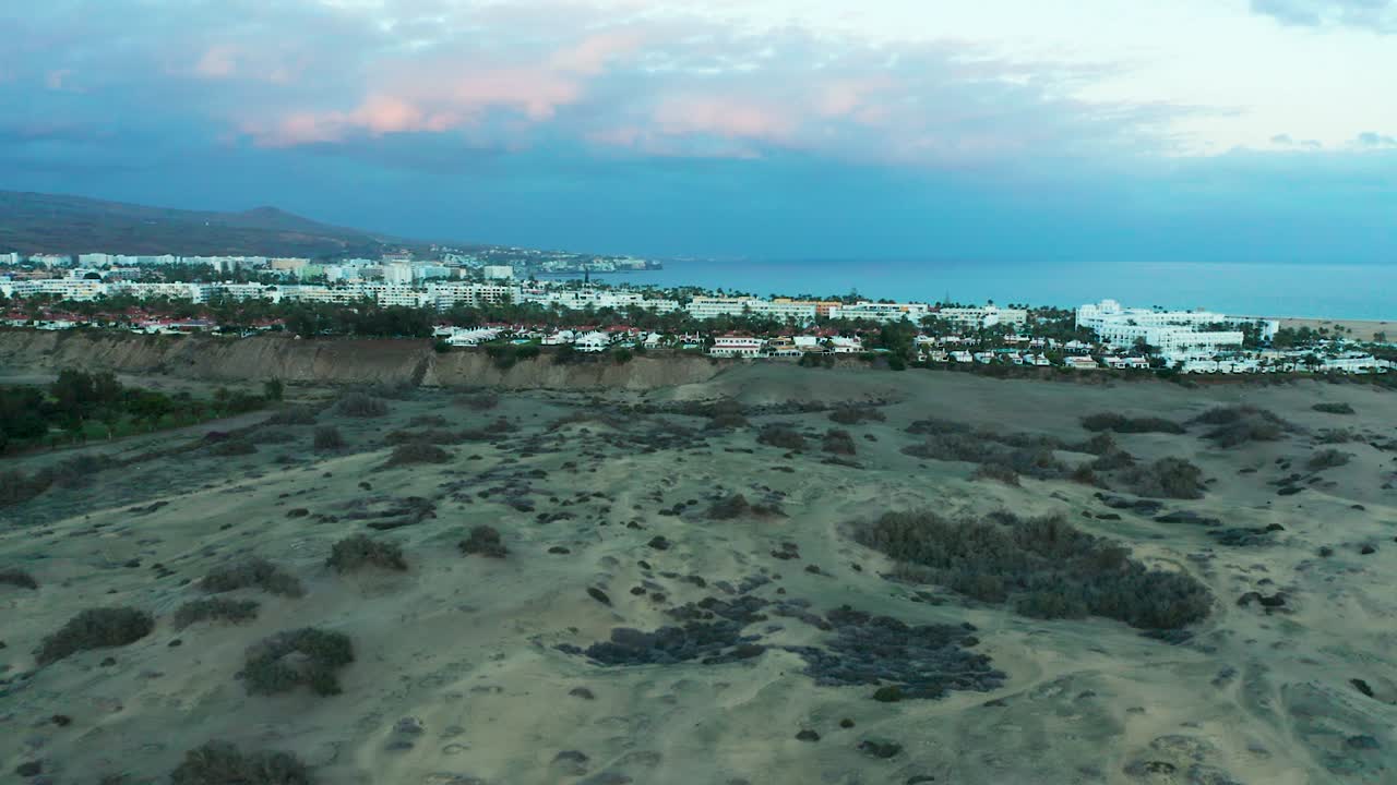Drone establishing overview of vast Maspalomas dunes with resorts looking out to sea, soft dawn light glows over sandy hills and expansive desert views