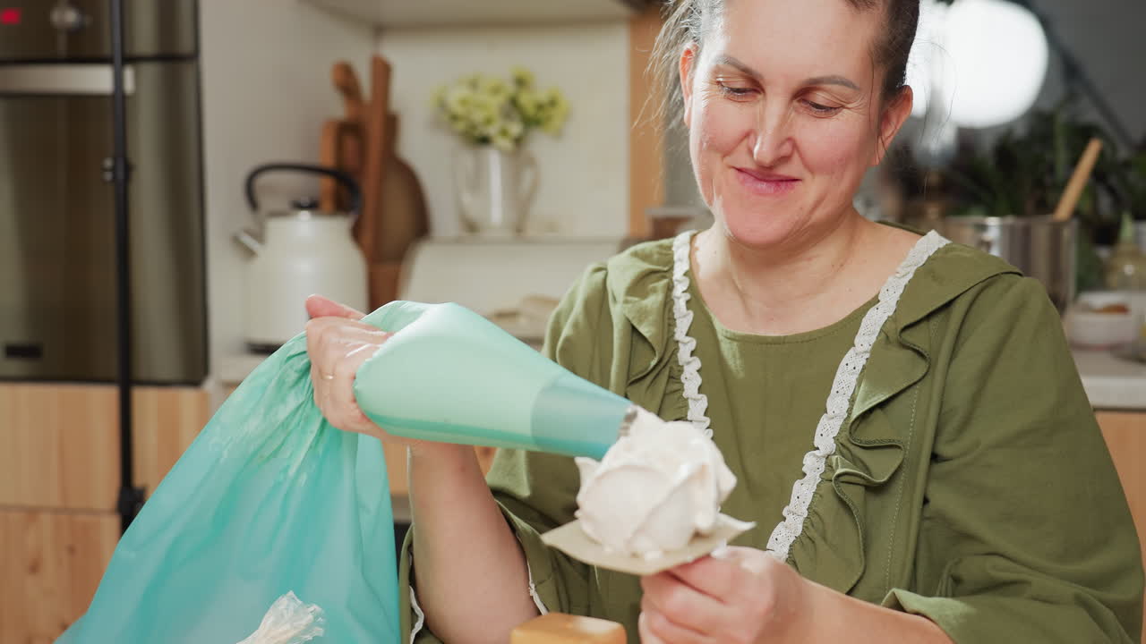 Joyful woman in green dress uses blue piping bag to apply thick white icing on dessert while smiling warmly in cozy kitchen, surrounded by baking tools