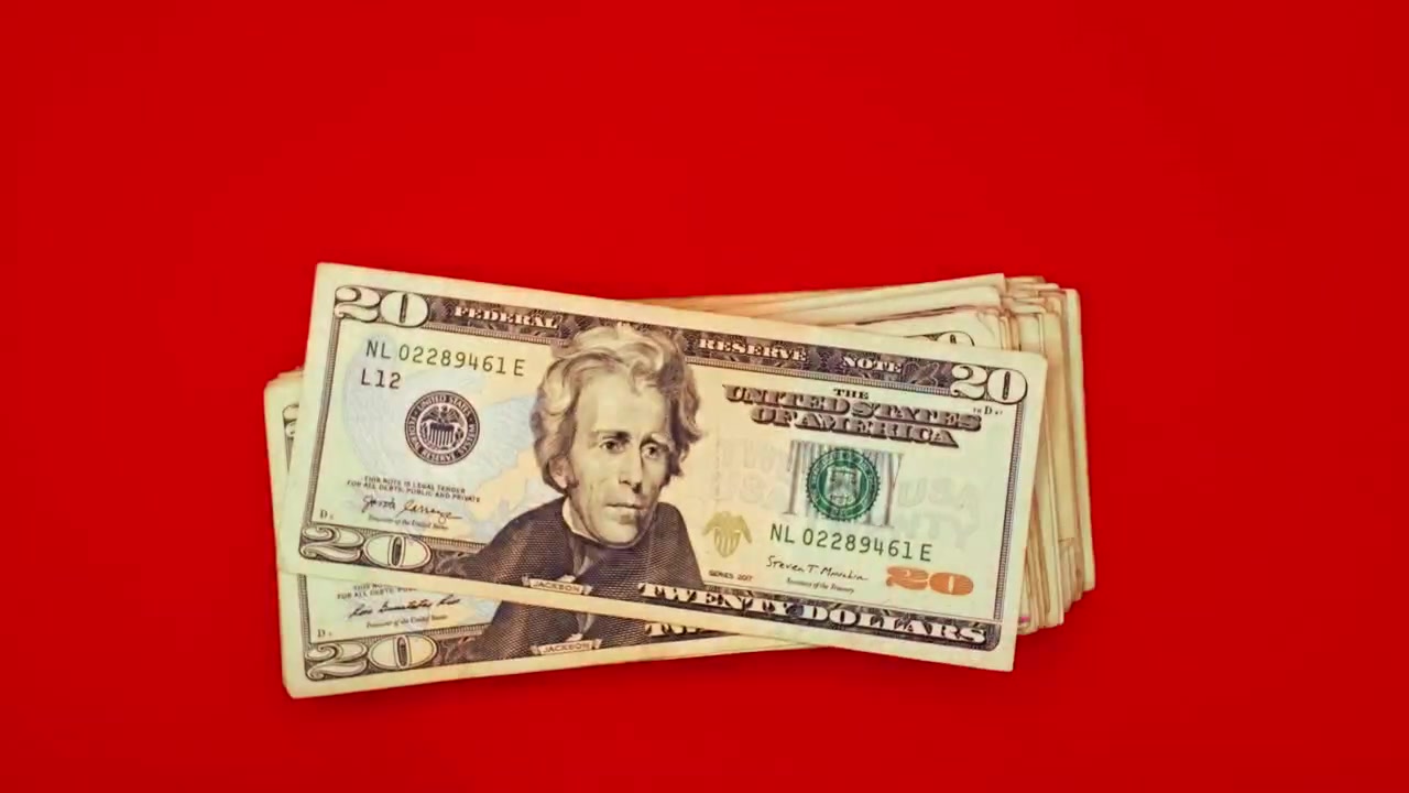 Nærbillede af en persons hænder, der tæller en vifte af tyve amerikanske dollar-sedler (USD) mod en ensfarvet rød baggrund. Koncept for kontant betaling, rigdom, ulovlig transaktion, finansiel succes eller lønudbetaling.
