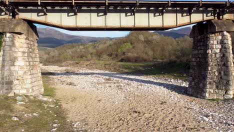 Luftaufnahmen,-Die-Zur-Goldenen-Stunde-Unter-Einer-Verlassenen-Eisenbahnstrecke-über-Einem-Ausgetrockneten-Flussbett-Vorbeifahren