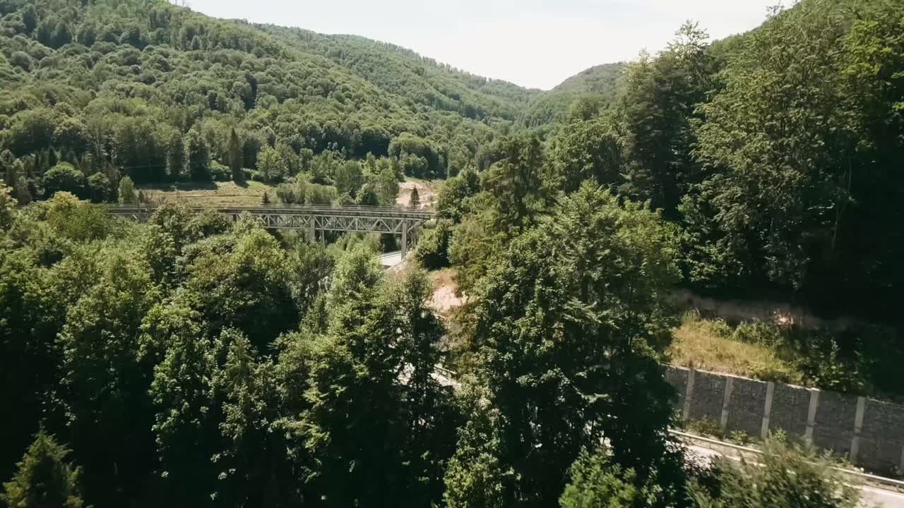 vista aérea dinámica con drones de carreras fpv volando sobre cabañas y bajo un viejo puente ferroviario junto a un camino sinuoso vacío