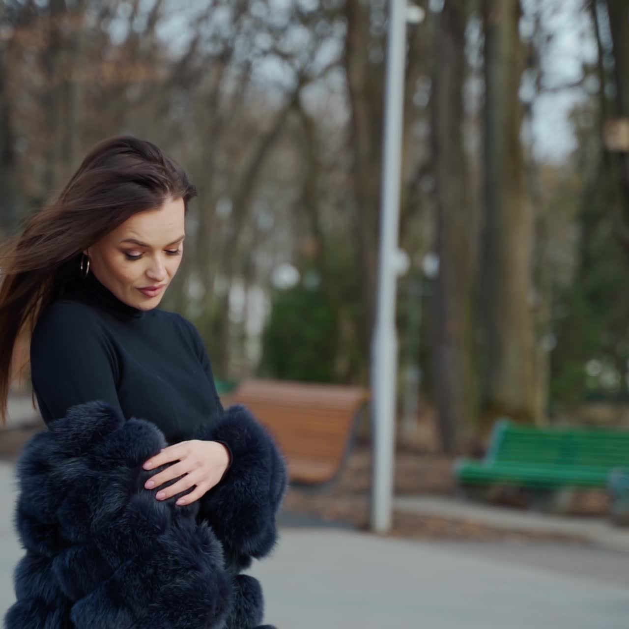 Fashionable girl with long hair looking at camera outdoors. Portrait of attractive young woman in fur coat poses on camera on the park background.