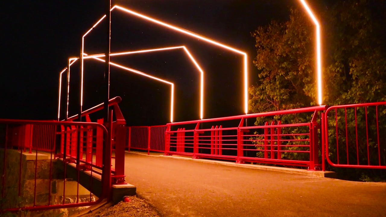 Imágenes en movimiento lento de un puente rojo metálico durante una noche de verano, bellamente iluminado por luces vanguardistas y artísticas. Algunos árboles son visibles en la noche oscura y el asfalto está iluminado por las luces LED