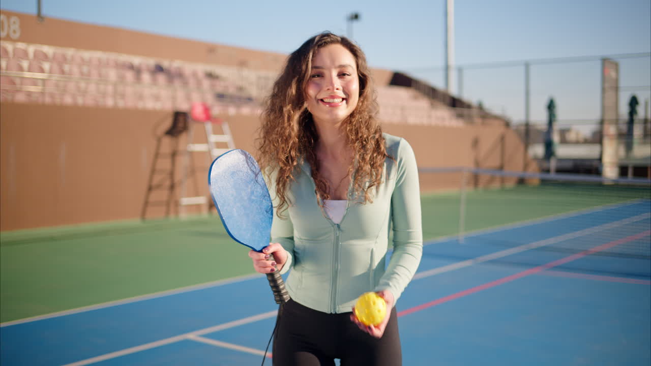A happy and smiling woman with curly hair holding a Pickleball blue racket and a yellow ball on a blue court on a sunny day