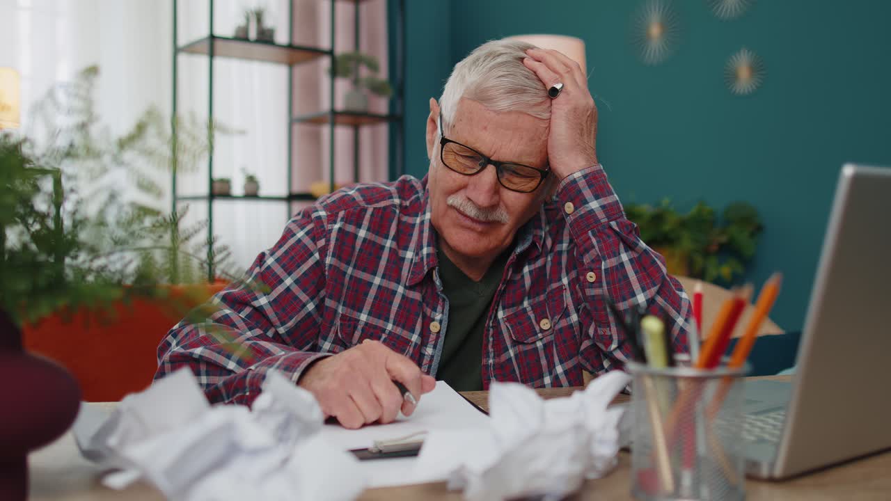 Senior man working on laptop computer making notes on sheet of paper upset by poor bad results