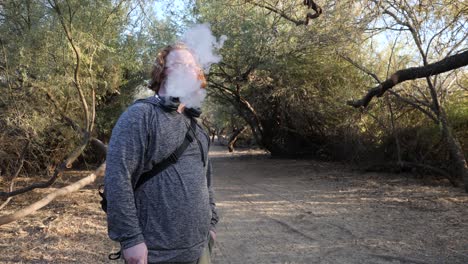 hombre de pelo rojo vaporizando de pie bajo los árboles.