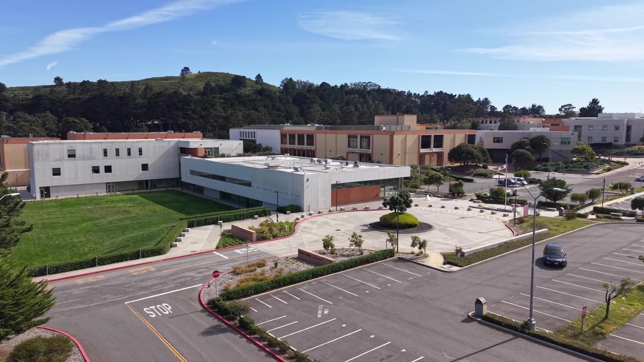 Vista aérea en una tarde soleada del campus de Skyline College, mostrando edificios modernos, una rotonda ajardinada, un estacionamiento vacío y las exuberantes colinas verdes de San Bruno al fondo.
