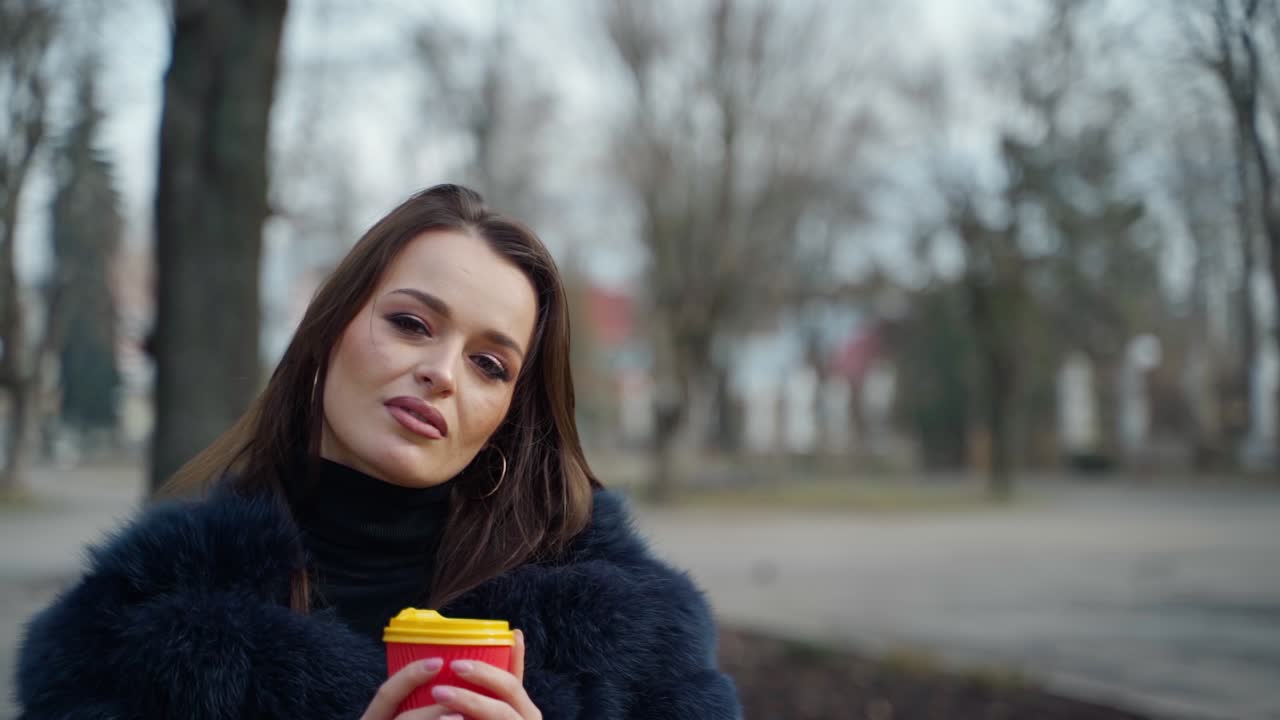 Attractive young woman with a plastic glass outdoors. Portrait of a charming girl with long dark hair in fur coat posing on camera in the park.