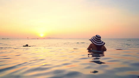 adult woman in swimsuit with hat in an infinity pool watching the sunset on the ocean and beach in the background