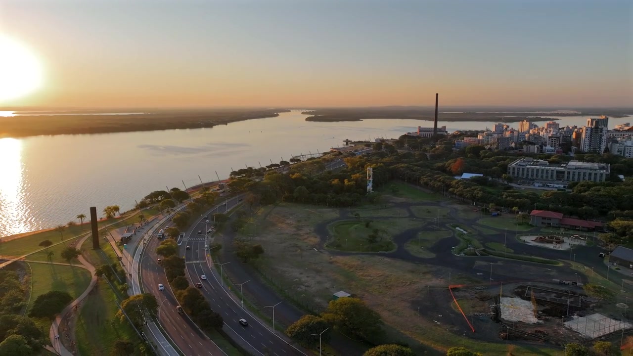 Strada curva lungo il fiume accanto a un parco con l'Usina do Gasômetro e lo skyline di Porto Alegre all'ora d'oro.