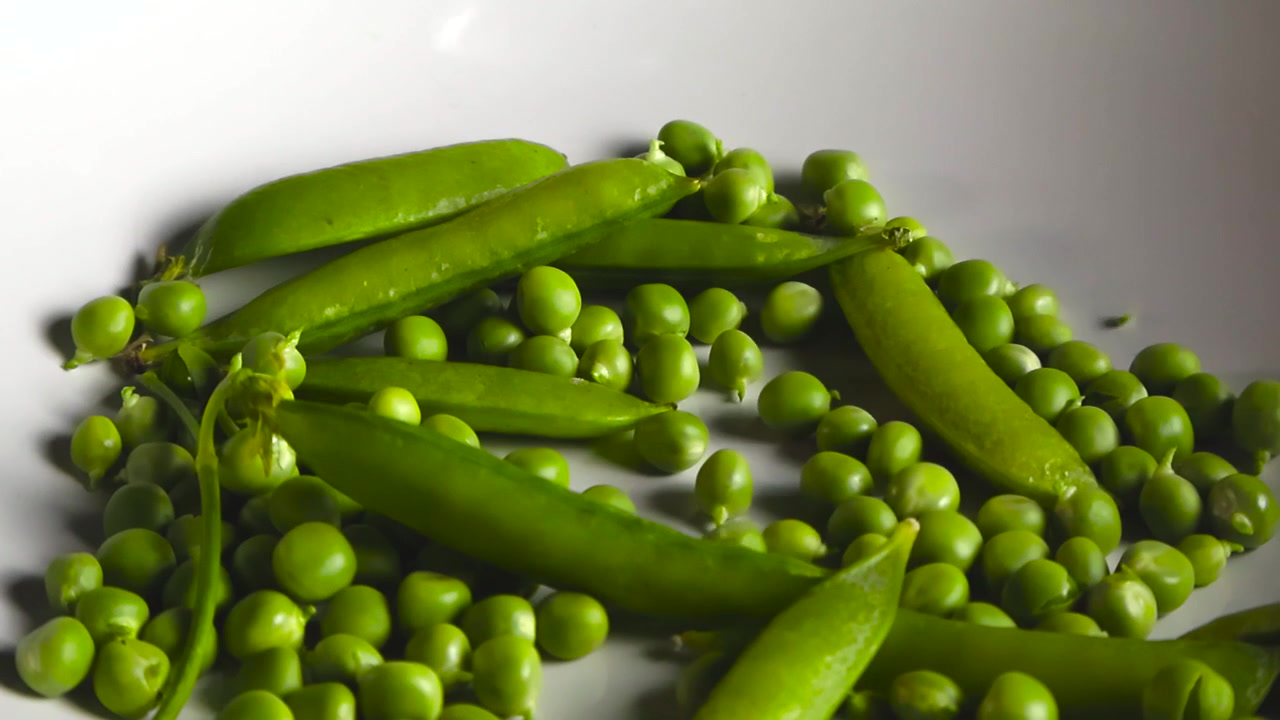 Numerosi piselli freschi e verdi che cadono al rallentatore in una ciotola bianca contenente diversi baccelli di piselli interi, creando un mucchio di verdure vivaci e salutari per un pasto nutriente