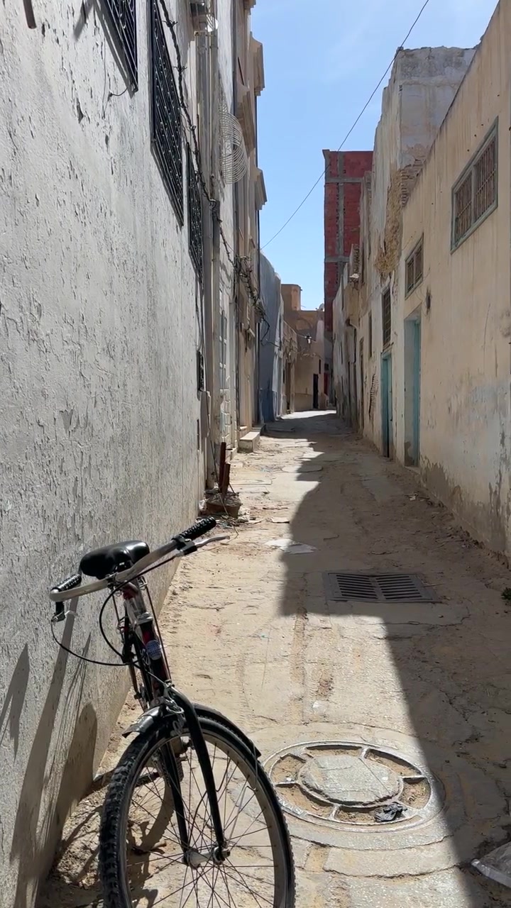 A lone bicycle leans against a worn wall in a narrow sunlit alley between old plaster buildings in Kairouan, Tunisia.