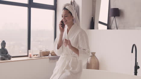 woman in bathrope with a towel in her head talking on the phone sitting on the edge of bathtub