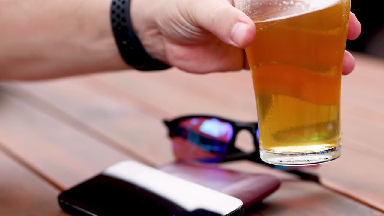 A hand reaches for a glass of beer beside sunglasses on a wooden table.
