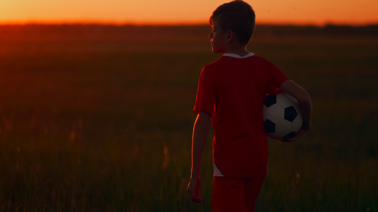 el niño está en el campo con la pelota en las manos mirando la puesta de sol y soñando con una carrera futbolística. la cámara sigue al niño