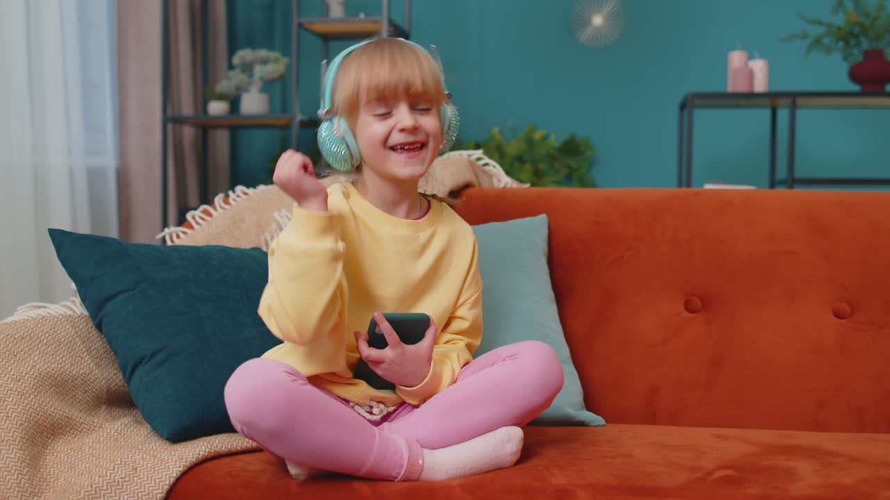 소파 위에서 혼자 집에서 음악을 듣는 동안 헤드폰을 착용한 스마트폰을 가진 소녀 아이가 춤을 추고 있습니다.
