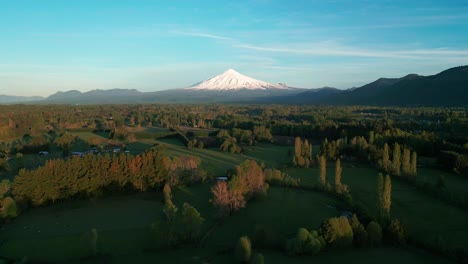 Reversing drone shot gliding away from a sunlit rural landscape toward a snow-capped Villarrica volcano under a clear blue sky at golden hour