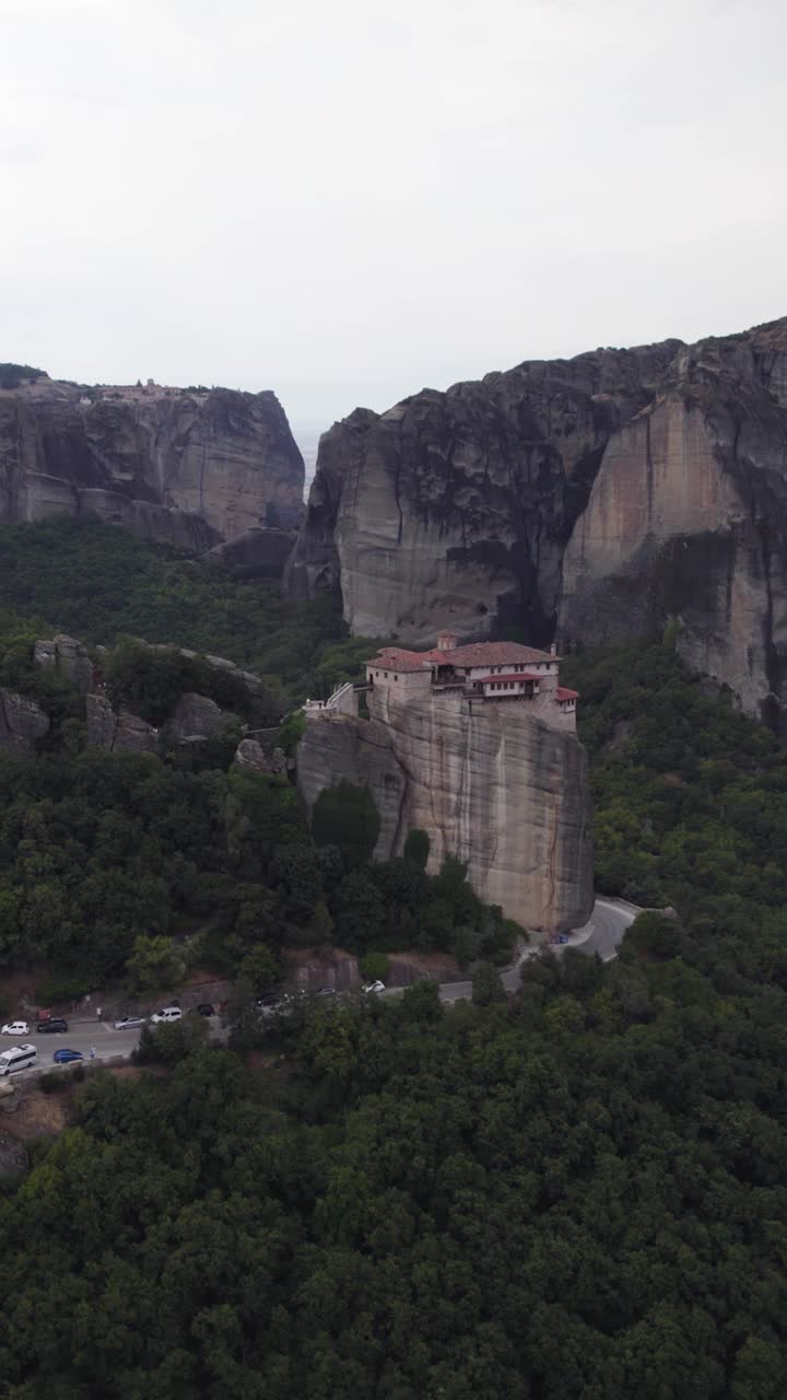 Vertical aerial of the Monastery of Rousanou perched atop a cliff in Meteora, Greece, showcasing the surrounding mountain landscape