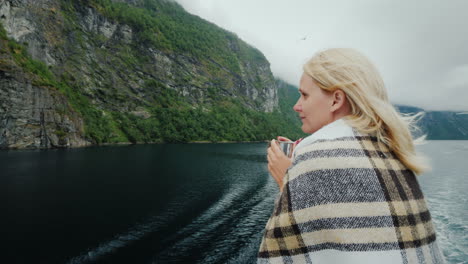 a woman with a cup of tea in her hands aboard a cruise ship looks at the beautiful mountains of the