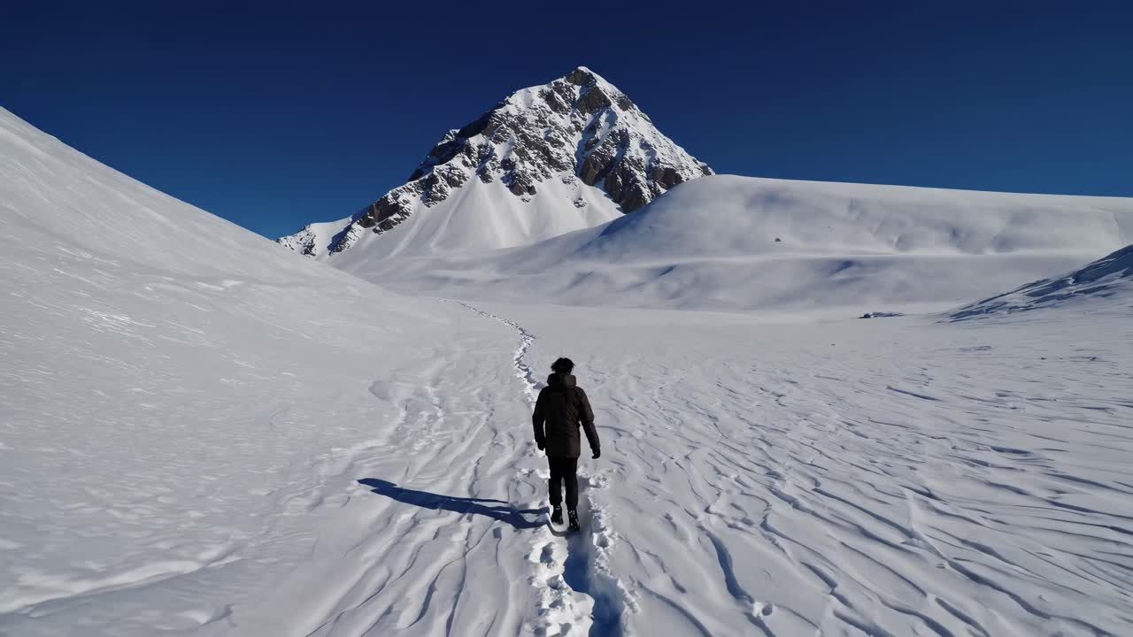 A person walks through a snowy landscape towards a mountain, captured from a low-angle