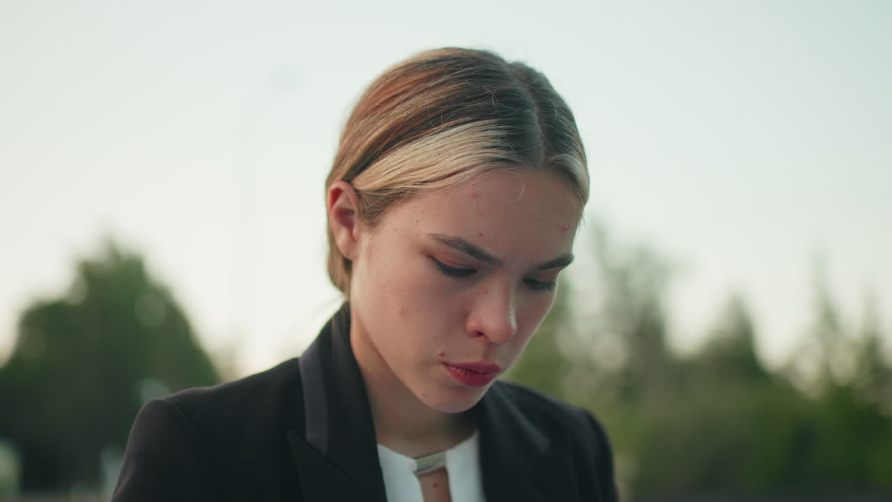 Close up of focused ceo with neat hairstyle intently reviewing work outdoors on wooden bench with blur background featuring greenery trees and building under clear sky
