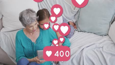 Animation-of-heart-icons-and-number-over-caucasian-grandmother-and-granddaughter-using-tablet