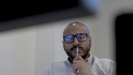 Bald-Ethnic-Minority-Male-Wearing-Glasses-In-Office-Swivelling-In-Chair-Tapping-Pen-On-His-Lips